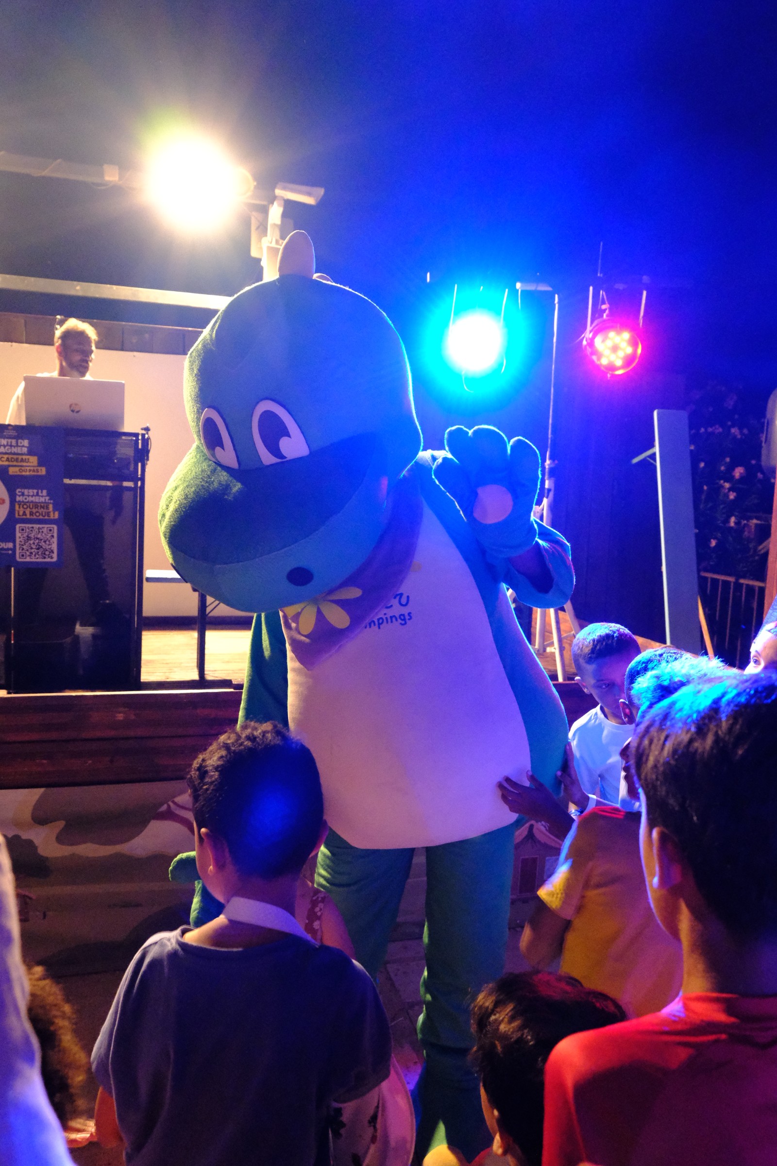 Soirée d'animation au camping avec mascotte et enfants devant scène éclairée.