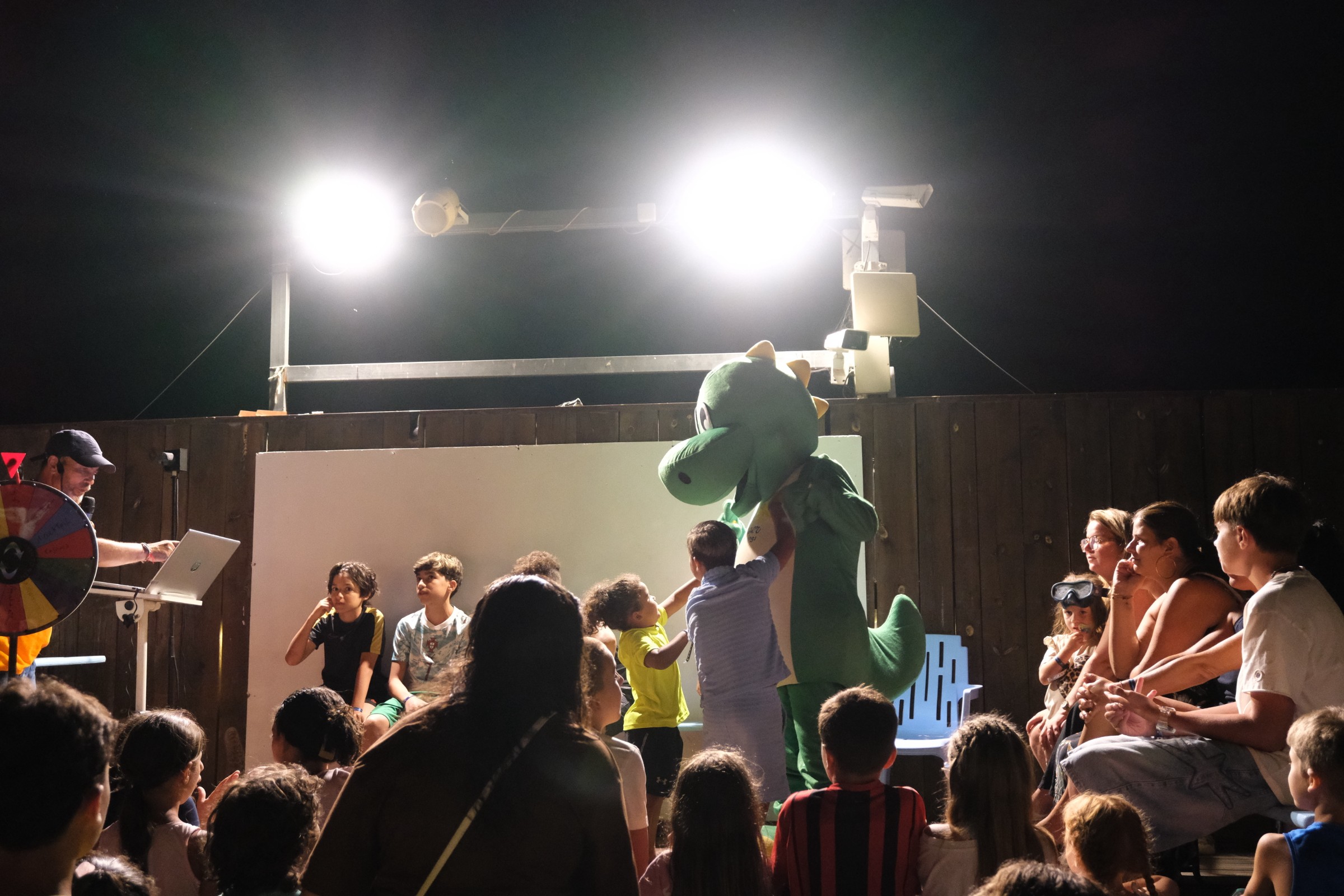 Spectacle de soirée avec mascotte verte sur scène et projecteurs illuminant l'assistance dans le camping.